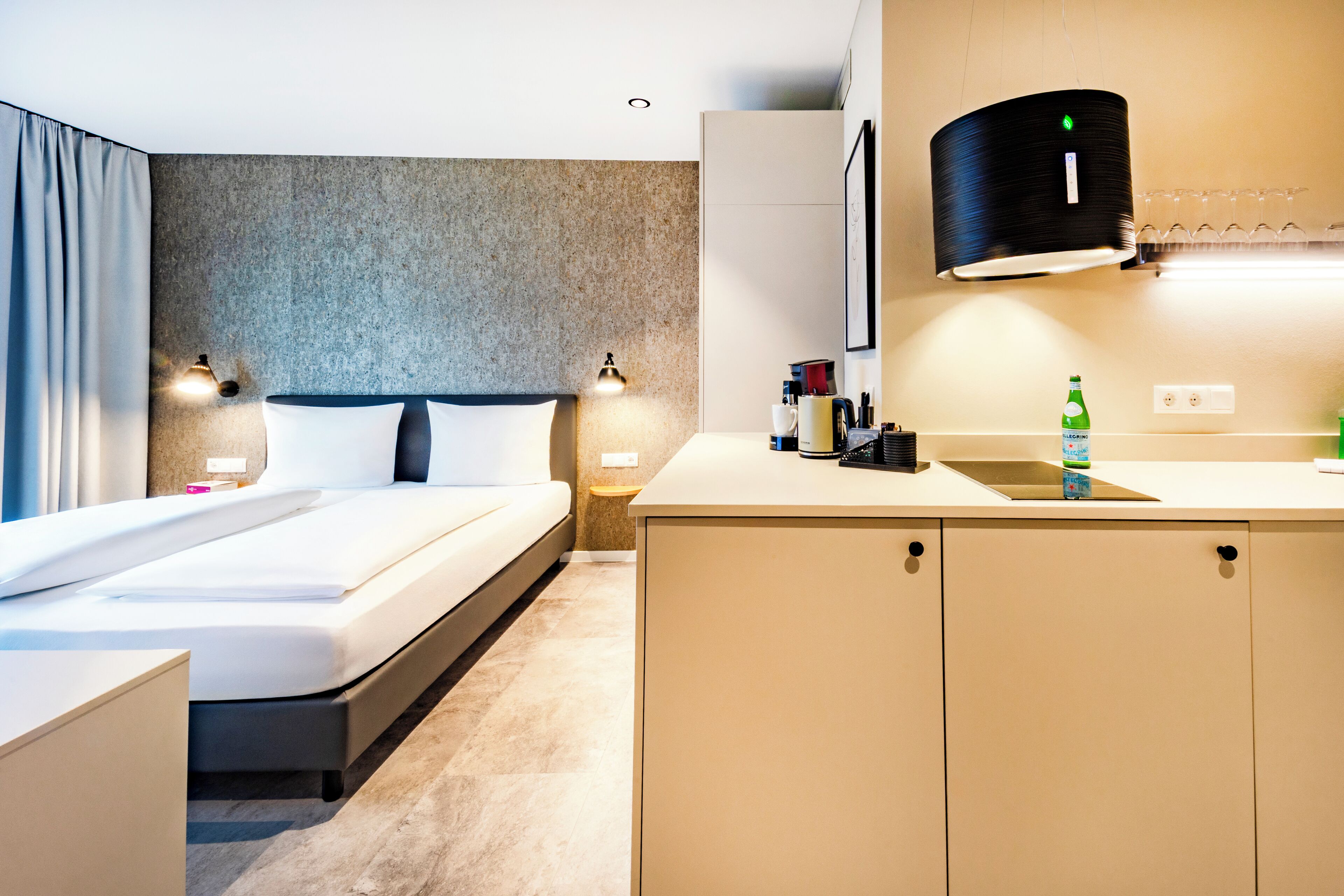A modern hotel room with a bed, bedside tables, a small kitchenette, and a large window.