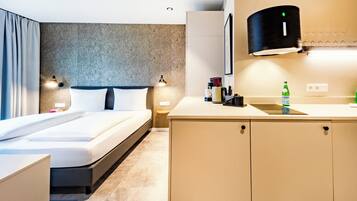 A modern hotel room with a bed, bedside tables, a small kitchenette, and a large window.