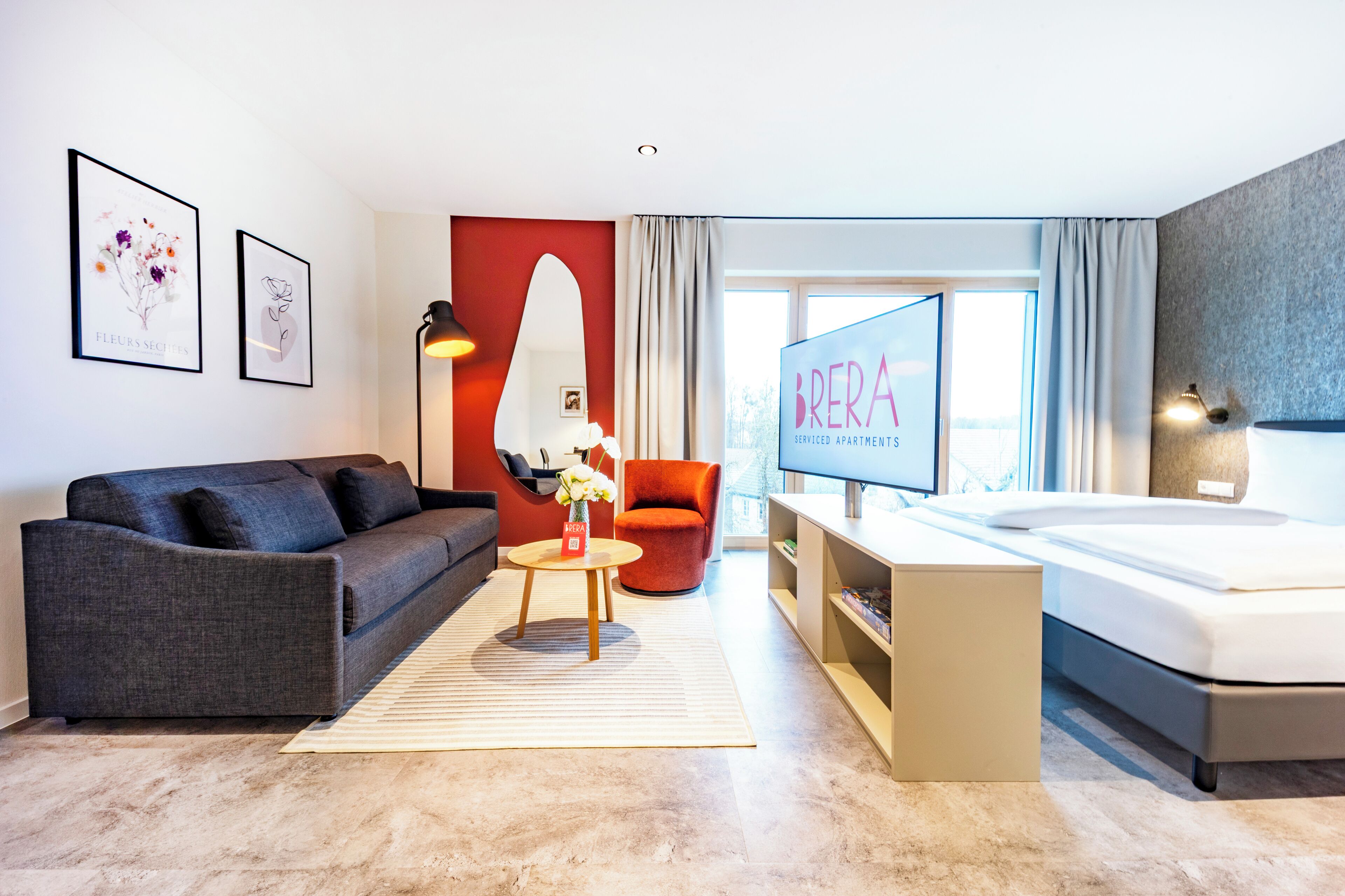 A modern hotel room with a grey sofa, a red armchair, a TV, a large bed, and a city view.