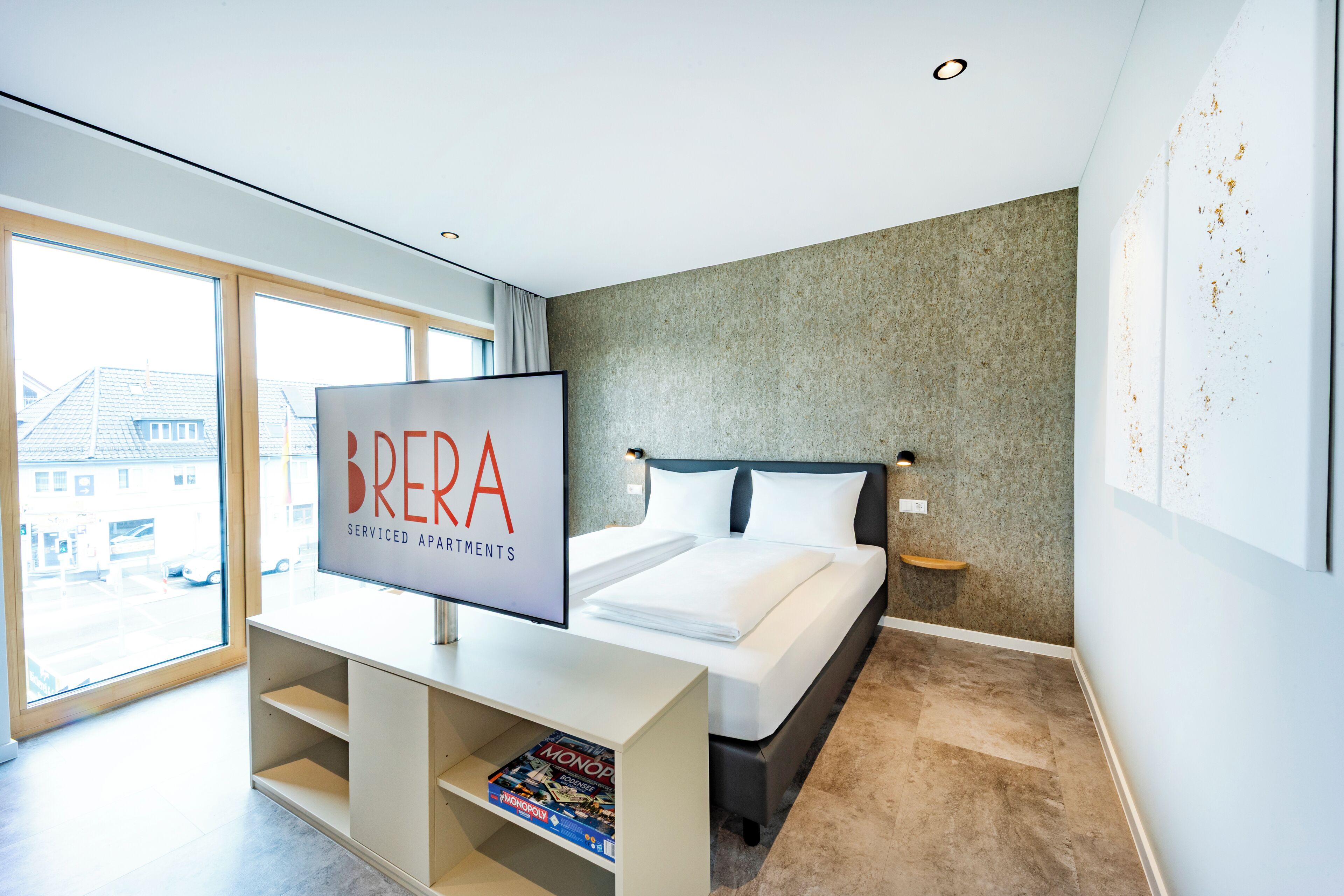 A modern bedroom with a large bed, a TV on a stand, and a window with a view of buildings outside.