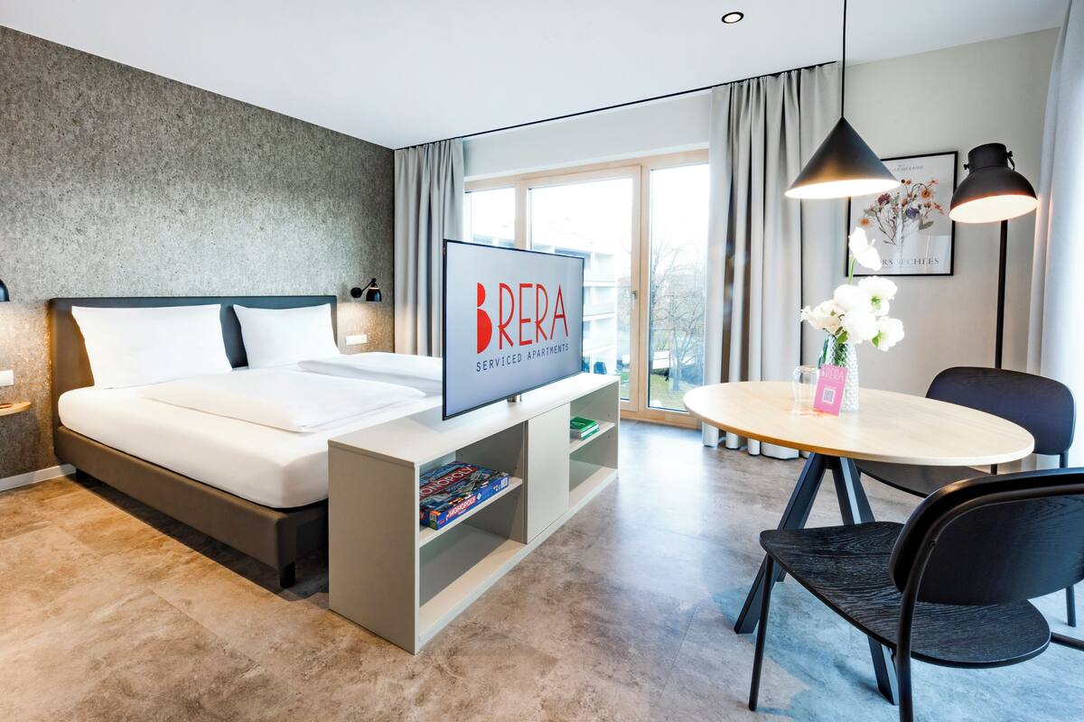 Ein modernes Hotelzimmer mit einem Bett, einem Fernseher, einem Essbereich mit Tisch und Stühlen sowie Blick ins Freie.