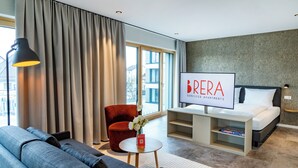 Una habitación de hotel moderna con un sofá, un sillón rojo, una mesita redonda con un florero, un televisor y una cama.