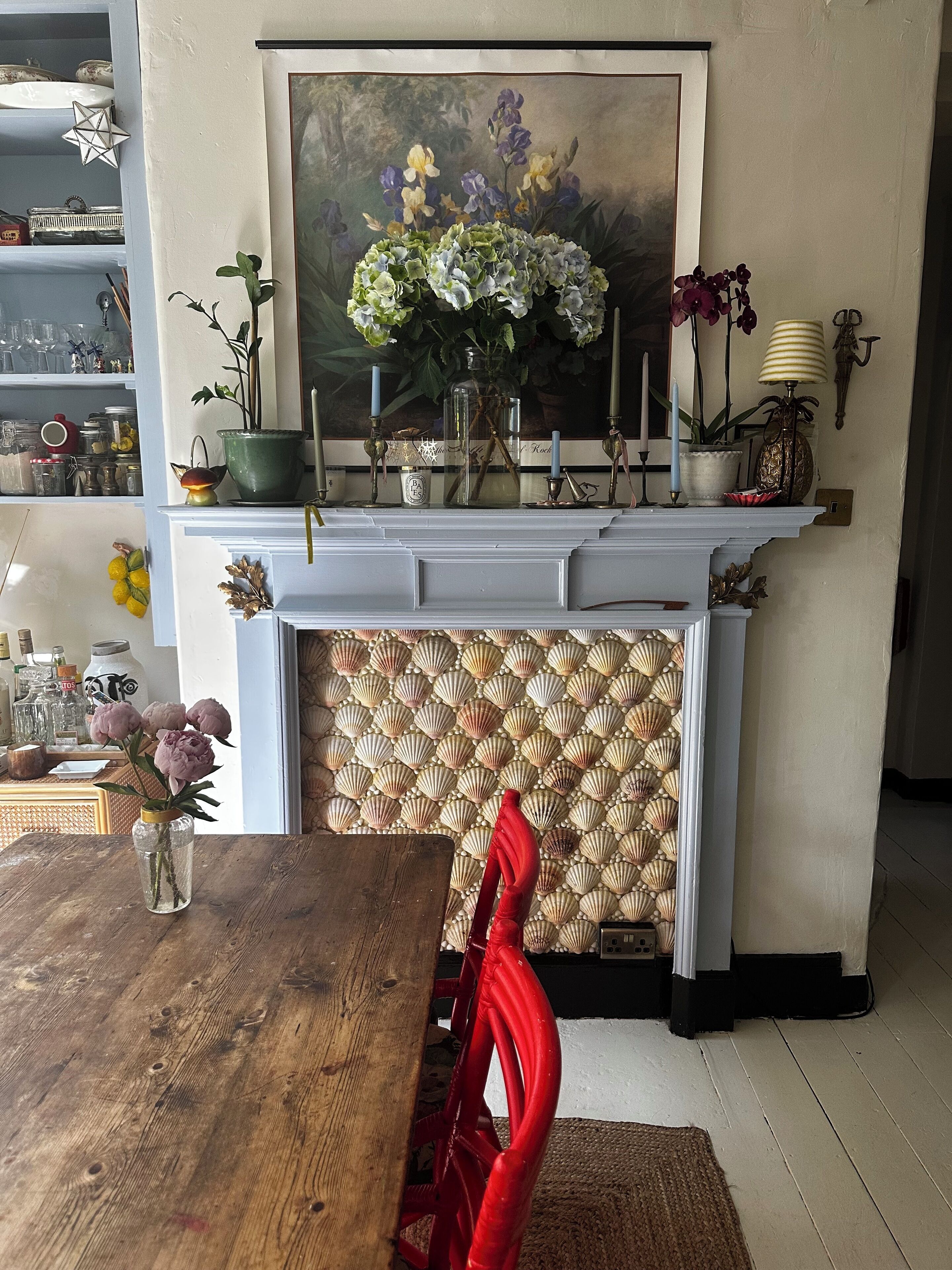 Un coin repas avec une table en bois, des chaises rouges, une cheminée ornée d’un manteau décoré, et un tableau accroché au-dessus du manteau.