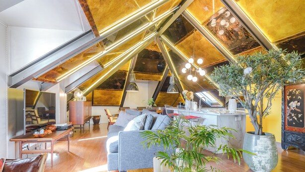 Een ruime loft met een hoge plafond, zichtbare balken, een centraal zithoek met een bank en salontafel, en een grote potplant.