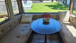 Een gezellige zithoek met een ronde tafel en kussens, een raam met uitzicht op een gazon en geparkeerde voertuigen.