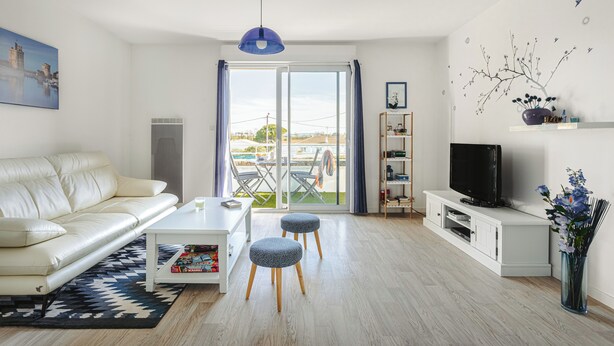 A modern living room with a sofa, ottomans, a coffee table, a TV on a white cabinet, and a view through a glass door.