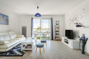 A modern living room with a sofa, ottomans, a coffee table, a TV on a white cabinet, and a view through a glass door. - Apartment in a secure complex, near La Rochelle, Châtelaillon-Plage (Aytré)