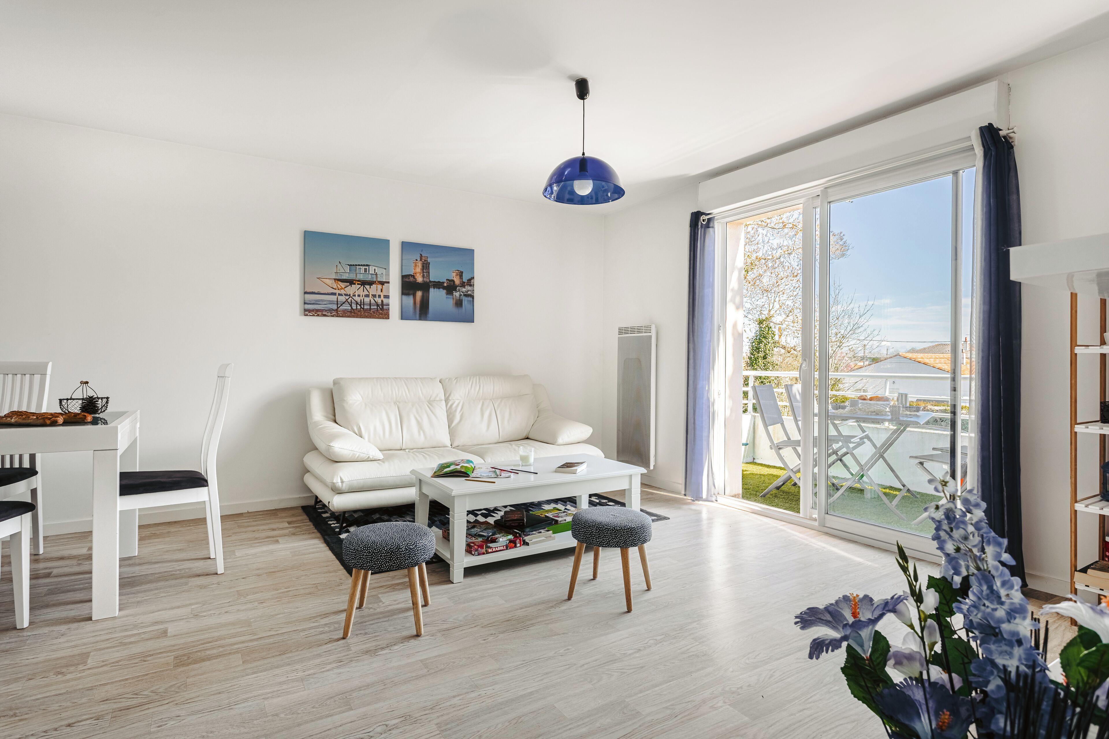 Een moderne woonkamer met een witte bank, een salontafel en twee bijpassende stoelen. Twee ingelijste foto's hangen aan de muur, er is een blauwe hanglamp en een groot raam met uitzicht op de omgeving.