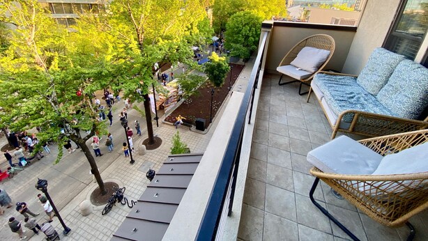 A balcony with wicker furniture, a tiled floor, and a view of a busy street with pedestrians and trees.