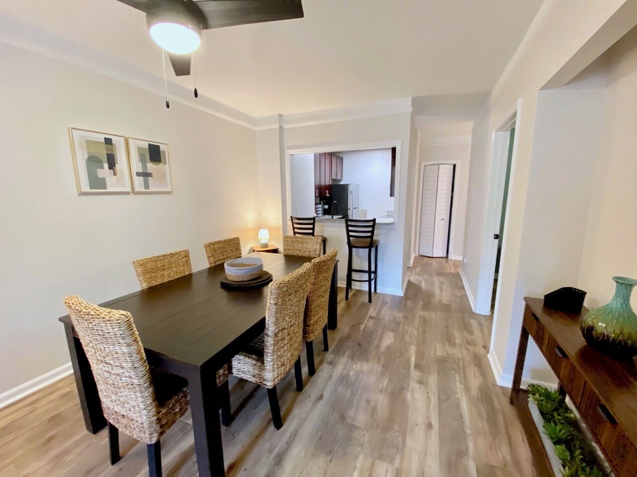 Un coin repas avec une table en bois foncé et des chaises en osier, une cuisine à l’arrière-plan et un couloir menant à une porte.