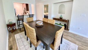 A dining area with a dark wooden table, woven chairs, a ceiling fan, and a kitchen in the background.