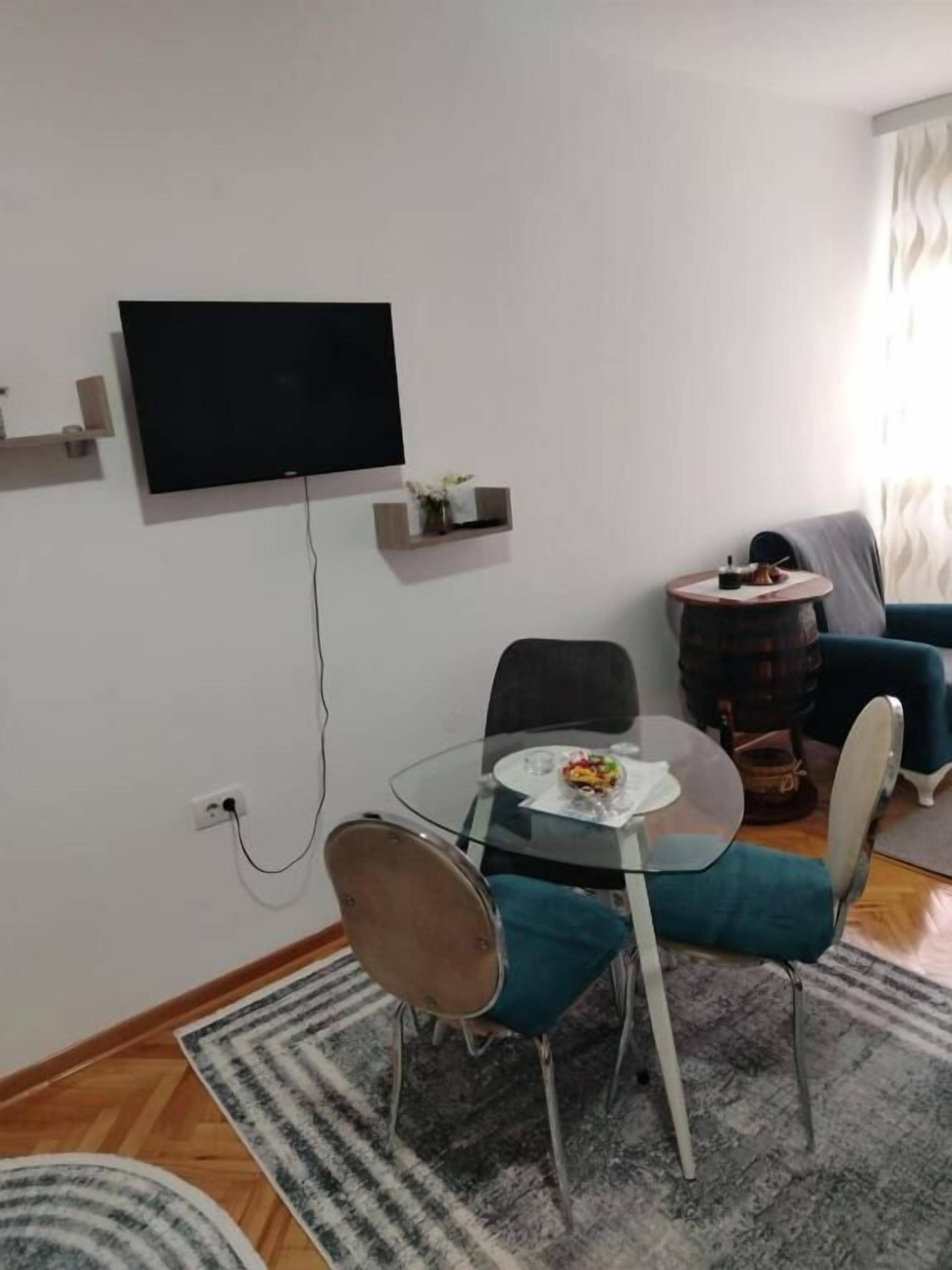 Ein modernes Wohnzimmer mit einem Glastisch, zwei Stühlen, einem flachbildfernseher an der Wand, einem kleinen Beistelltisch und einem blauen Sofa.
