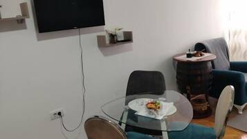 Ein modernes Wohnzimmer mit einem Glastisch, zwei Stühlen, einem flachbildfernseher an der Wand, einem kleinen Beistelltisch und einem blauen Sofa.