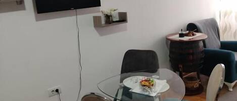 Un salon moderne avec une table à manger en verre, deux chaises, un téléviseur à écran plat fixé au mur, une petite table d’appoint et un canapé bleu.
