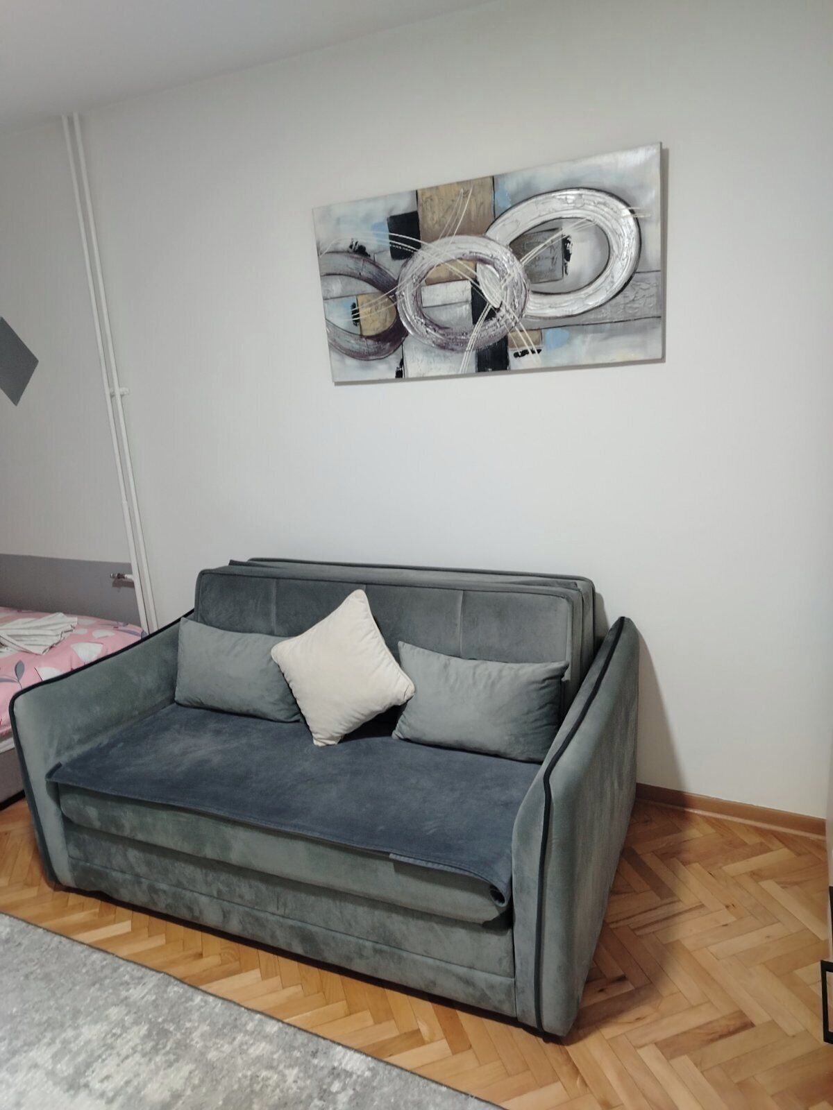 A grey sofa with cushions, a wooden herringbone floor, and a wall-mounted abstract art piece.