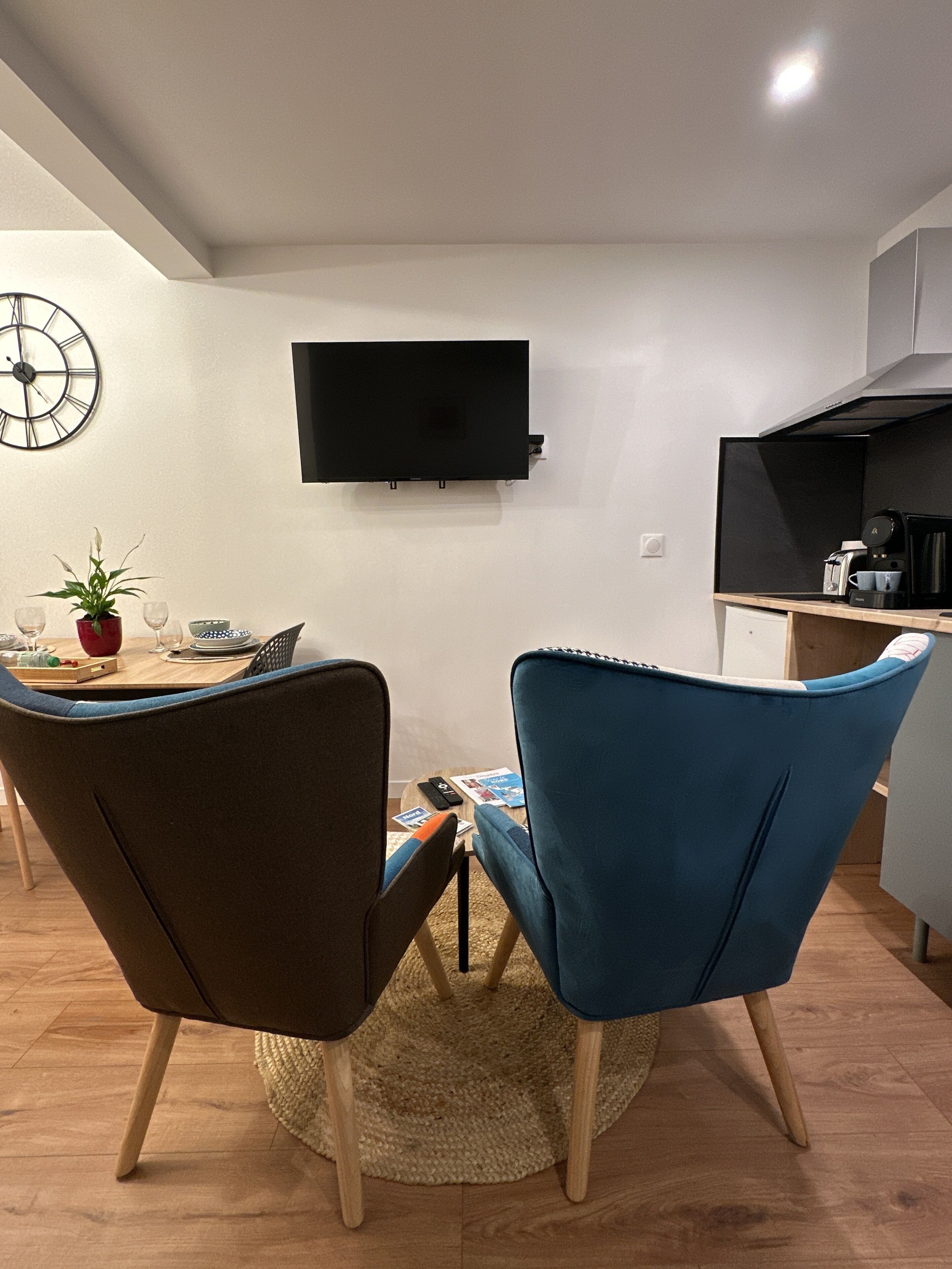 Een moderne woonkamer met een flatscreen televisie aan de muur, een eettafel met stoelen, een koffieapparaat en een klok.