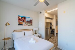 Hotellihuone, jossa on sänky, yöpöytä, lamppu, aavikkomaiseman kuva ja näkymä keittiötilaan.