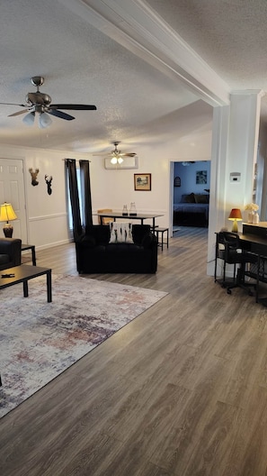 A spacious living room with a black sofa, a coffee table, a dining table, and a ceiling fan.