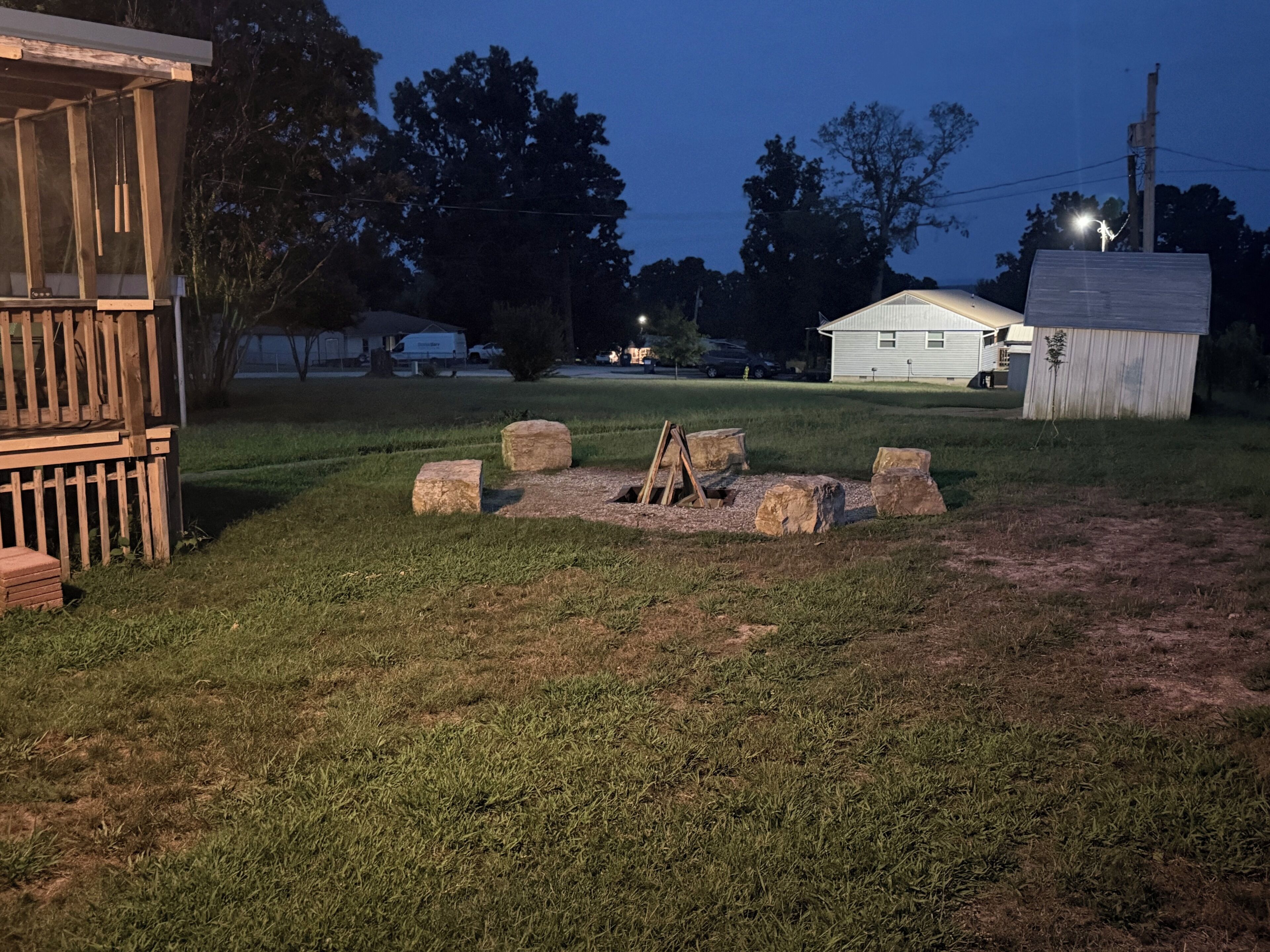 A grassy outdoor area with a wooden structure, a stone fire pit, and a small shed.