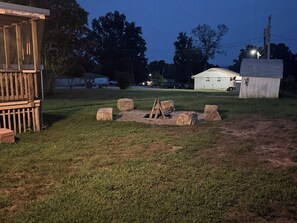 A grassy outdoor area with a wooden structure, a stone fire pit, and a small shed.