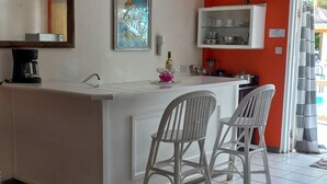 Una cocina con encimera blanca, dos sillas blancas, cafetera y un armario con vajilla.