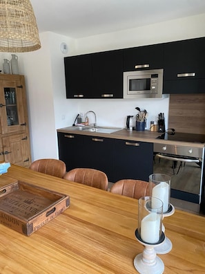Eine moderne Küche mit schwarzen Schränken, einer Mikrowelle aus Edelstahl und einem Backofen. Ein Holztisch mit braunen Stühlen und eine Flasche Champagner sind ebenfalls vorhanden.