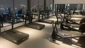 Une salle de sport équipée de tapis de course et de vélos d’exercice, de grandes fenêtres offrant une vue sur la ville et un aménagement intérieur moderne.