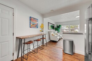Eine Küche mit einer Holztresen, drei Barhocker, einem Mülleimer, einem weißen Sofa und einem Fenster mit Blick auf Häuser.