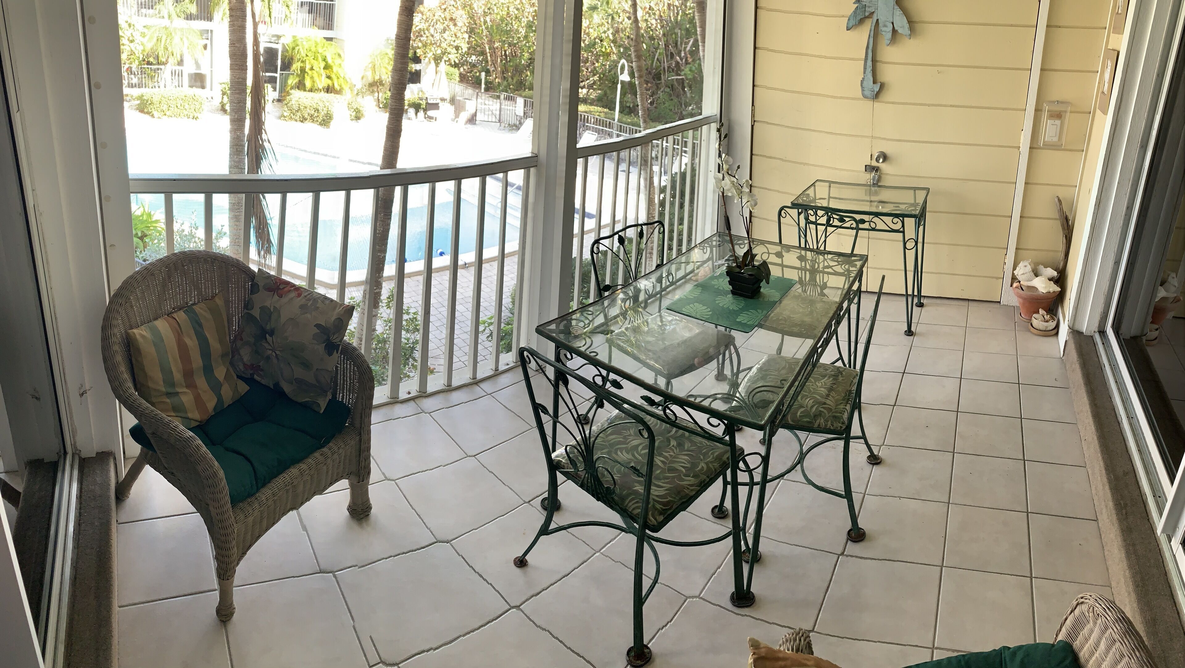 A balcony with a wicker chair, a glass table, and chairs, overlooking a pool and greenery.