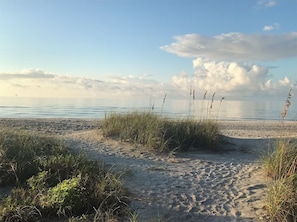 Ein Strand mit Sand, Gras und ruhiger See unter einem teils bewölkten Himmel.