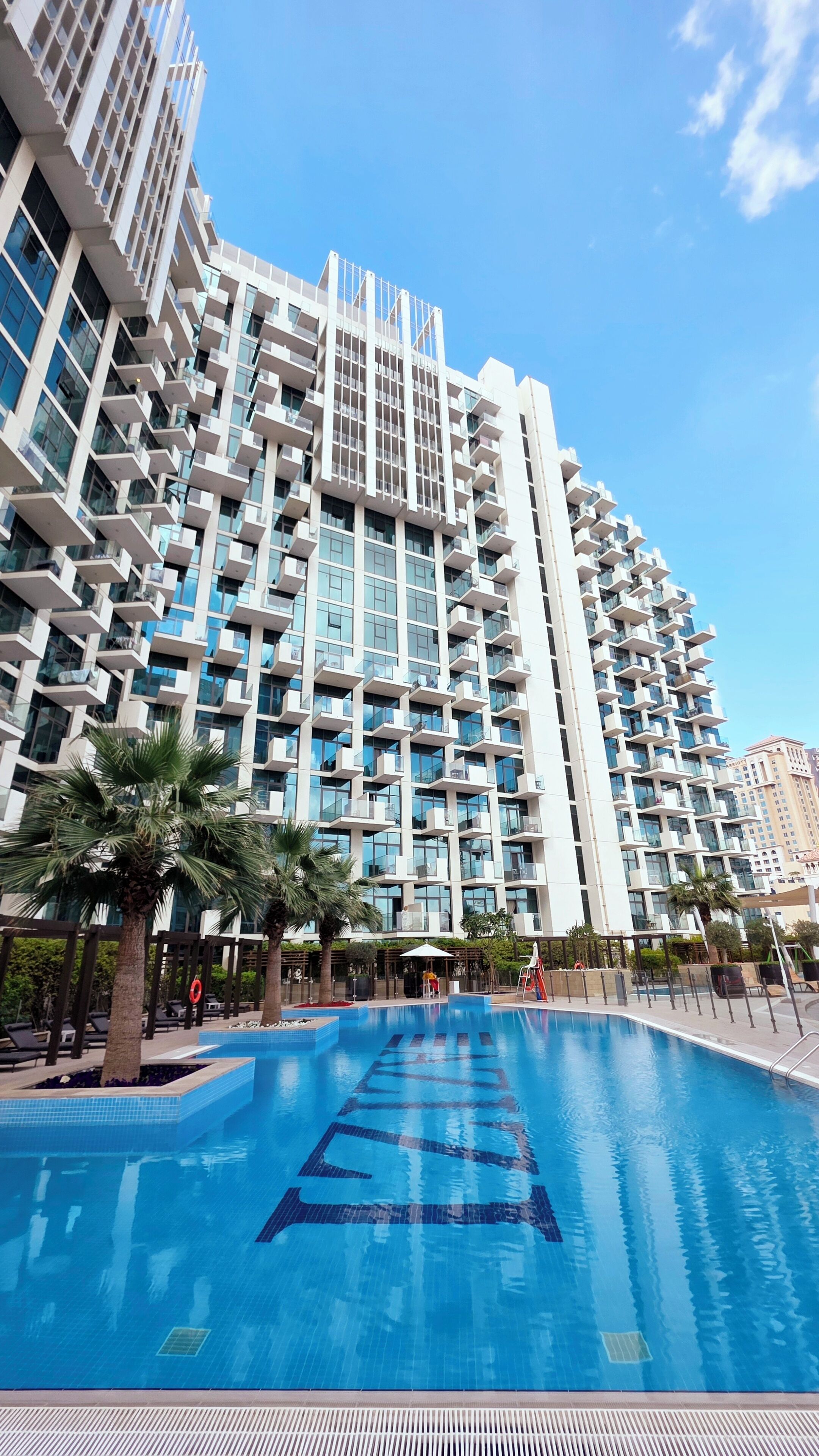 A modern hotel with a large pool area, featuring a unique architectural design with balconies and a clear blue sky.