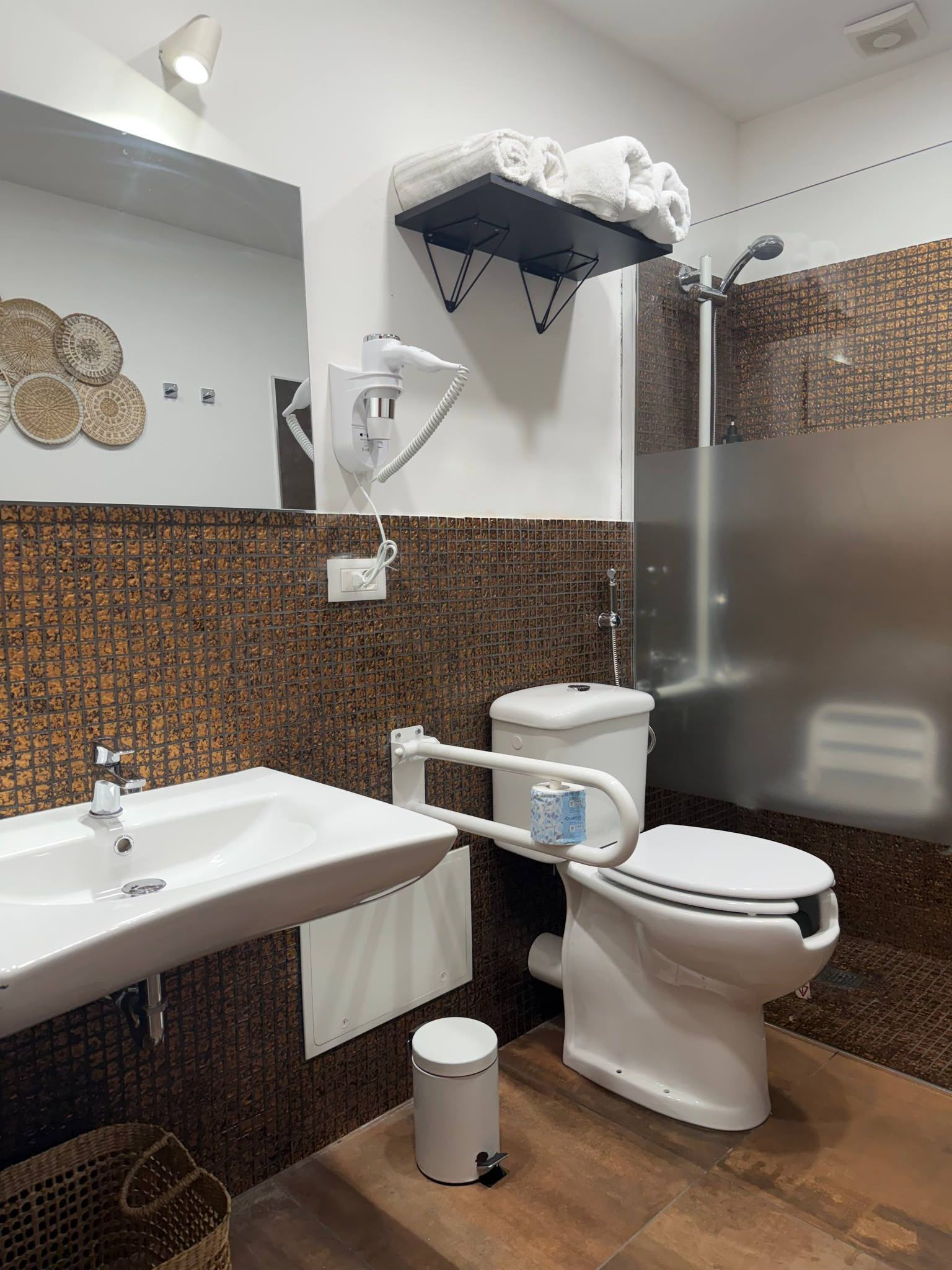 Une salle de bain avec un lavabo, des toilettes et une douche, le tout de couleur blanche. On trouve un porte-serviettes au-dessus des toilettes et un luminaire fixé au mur.