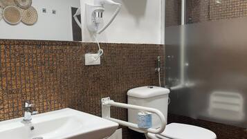 Une salle de bain avec un lavabo, des toilettes et une douche, le tout de couleur blanche. On trouve un porte-serviettes au-dessus des toilettes et un luminaire fixé au mur.