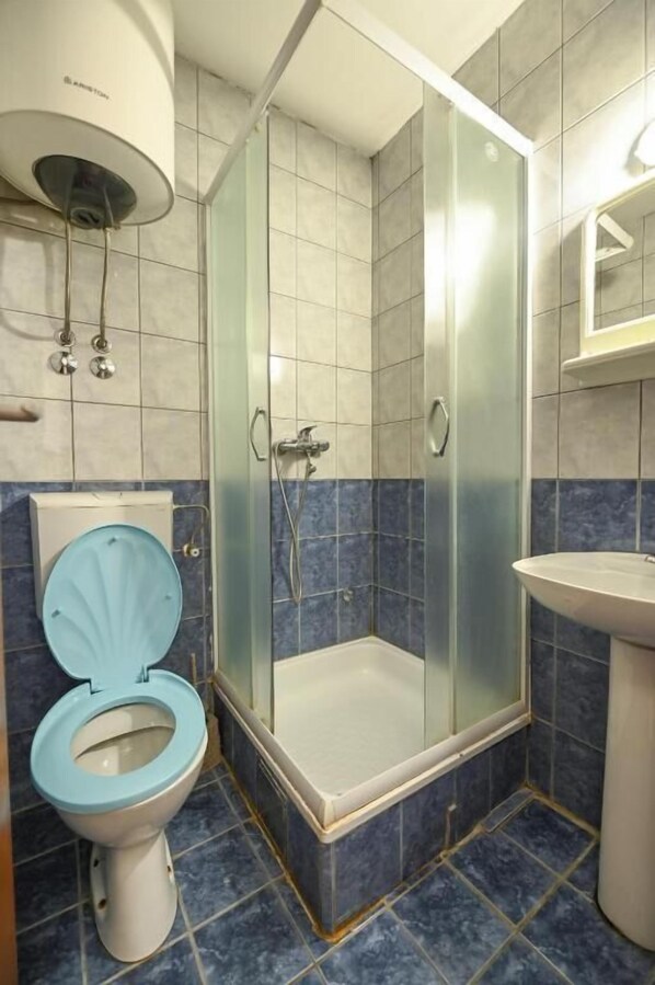 A bathroom with a glass-enclosed shower, a toilet with a blue seat, and a sink. - HOSTEL IZVOR (Podgorica)