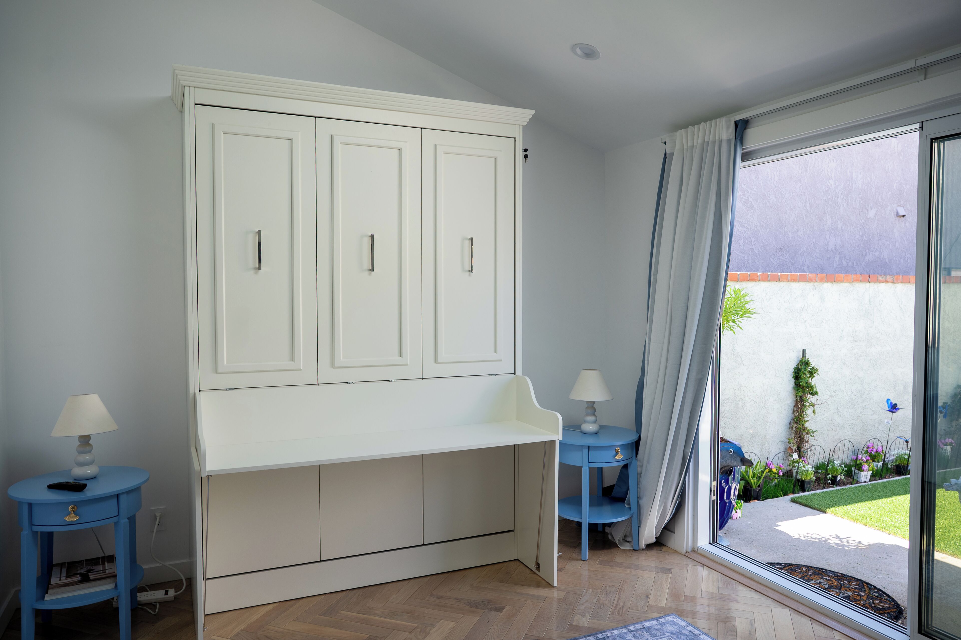 Une pièce avec un meuble blanc, deux petites tables avec des lampes et une grande fenêtre avec des rideaux.
