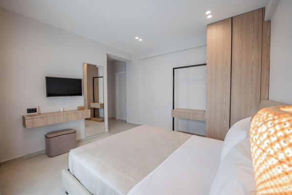 A modern hotel room with a large bed, a flat-screen TV mounted on the wall, a wooden wardrobe, and a small trash bin.
