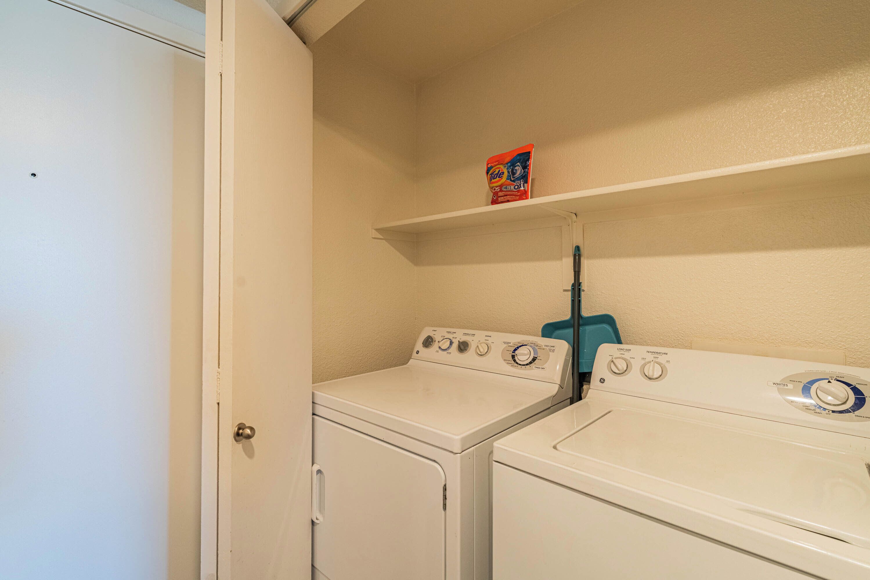 A laundry area with two white washers and dryers, a shelf with a detergent box, and a door to the left.