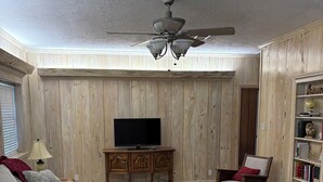 Un salón con televisor, paneles de madera, ventilador de techo, mecedora y estantería.