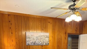 Un dormitorio con pared de paneles de madera, una cama con colcha azul, una mesita de noche con lámpara y un ventilador de techo.