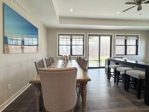 A dining area with a large table and chairs, a kitchen area with bar stools, and large windows with a view of the outdoors. - Fairways Townhome | Birchwood Golf & CC (Harbor Springs)