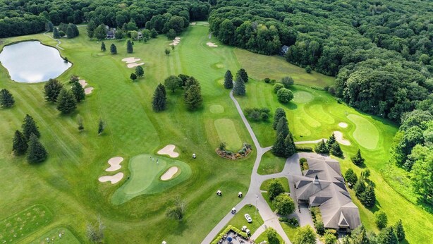 Luftaufnahme eines Golfplatzes mit einem See, umgeben von dichtem Wald und einem großen Gebäude mit markanter Dachform.