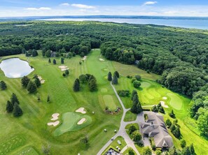 Oversigtsbillede af en golfbane med en sø, omgivet af tæt skov, og en stor bygning med et markant tag.