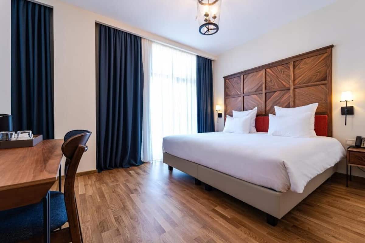 A hotel room with a large bed, a wooden headboard, a desk with a chair, and a window with curtains.