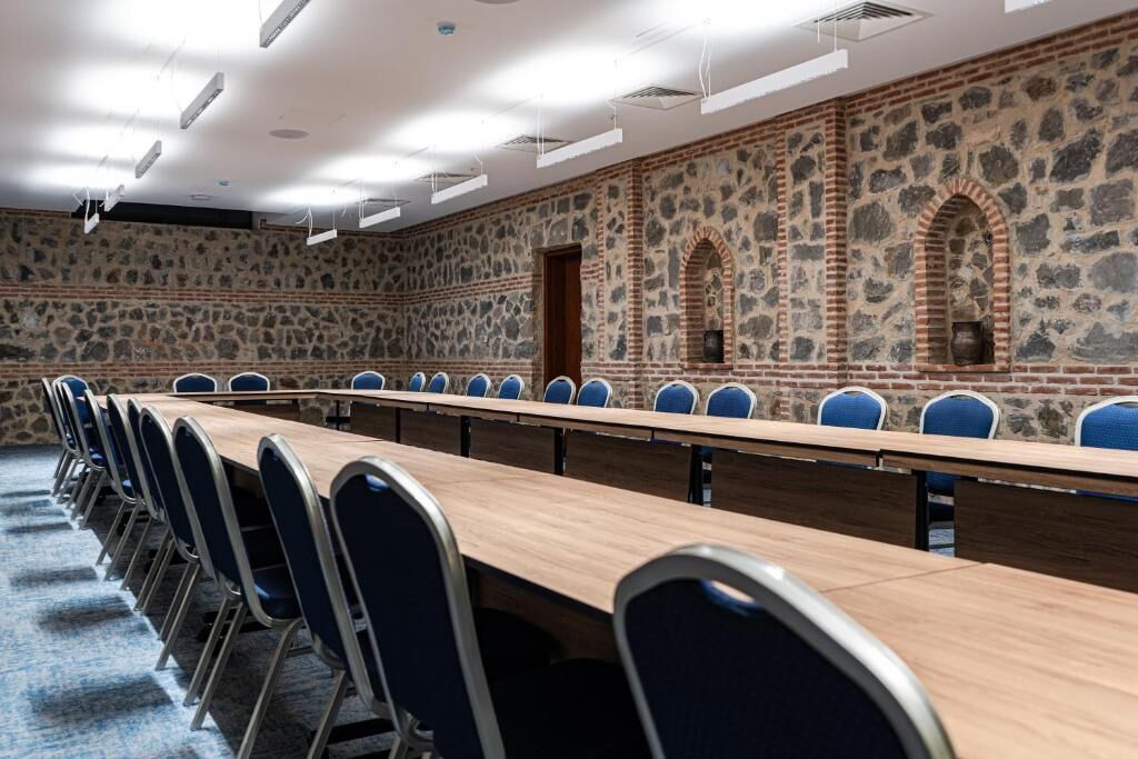 Un salón de conferencias con una larga mesa y sillas azules, paredes de piedra y una puerta de madera.