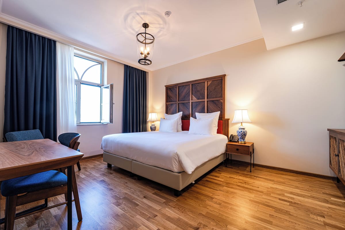 A hotel room with a bed, bedside table, lamp, window with curtains, and a wooden floor.