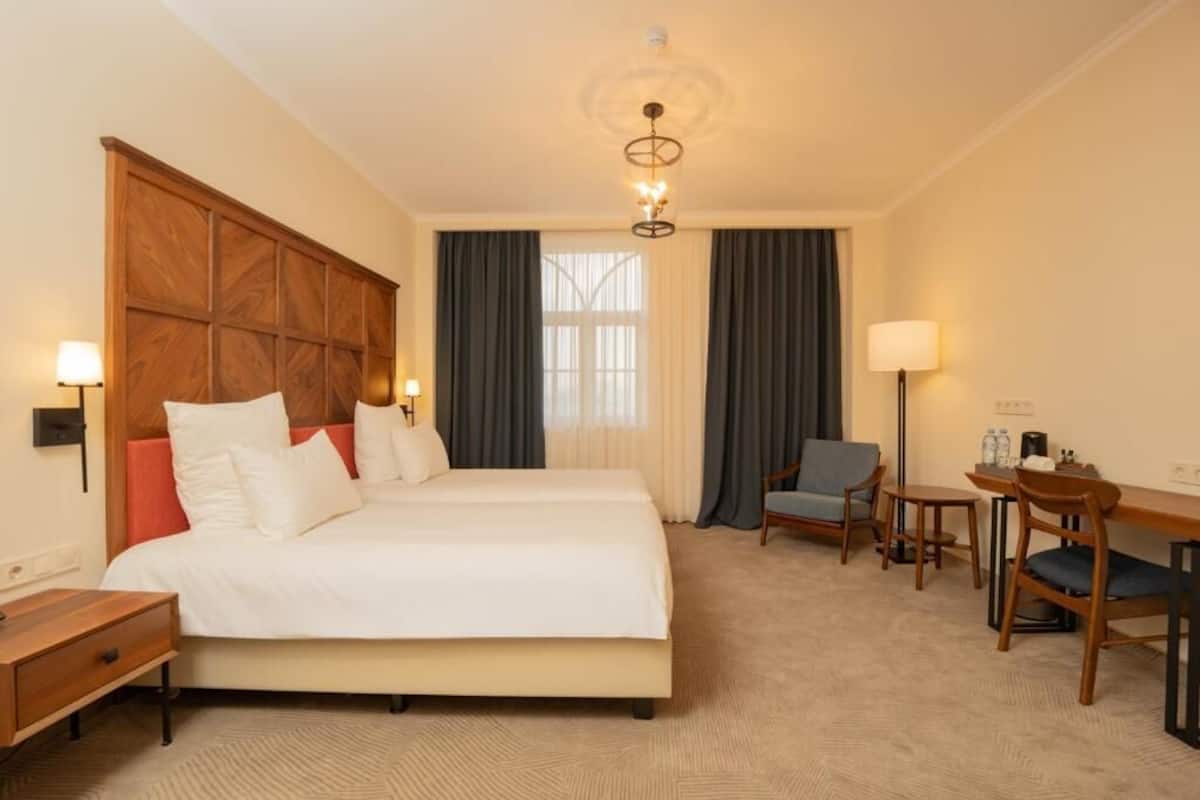 A hotel room with a large bed, a desk, two chairs, a nightstand, and a chandelier.