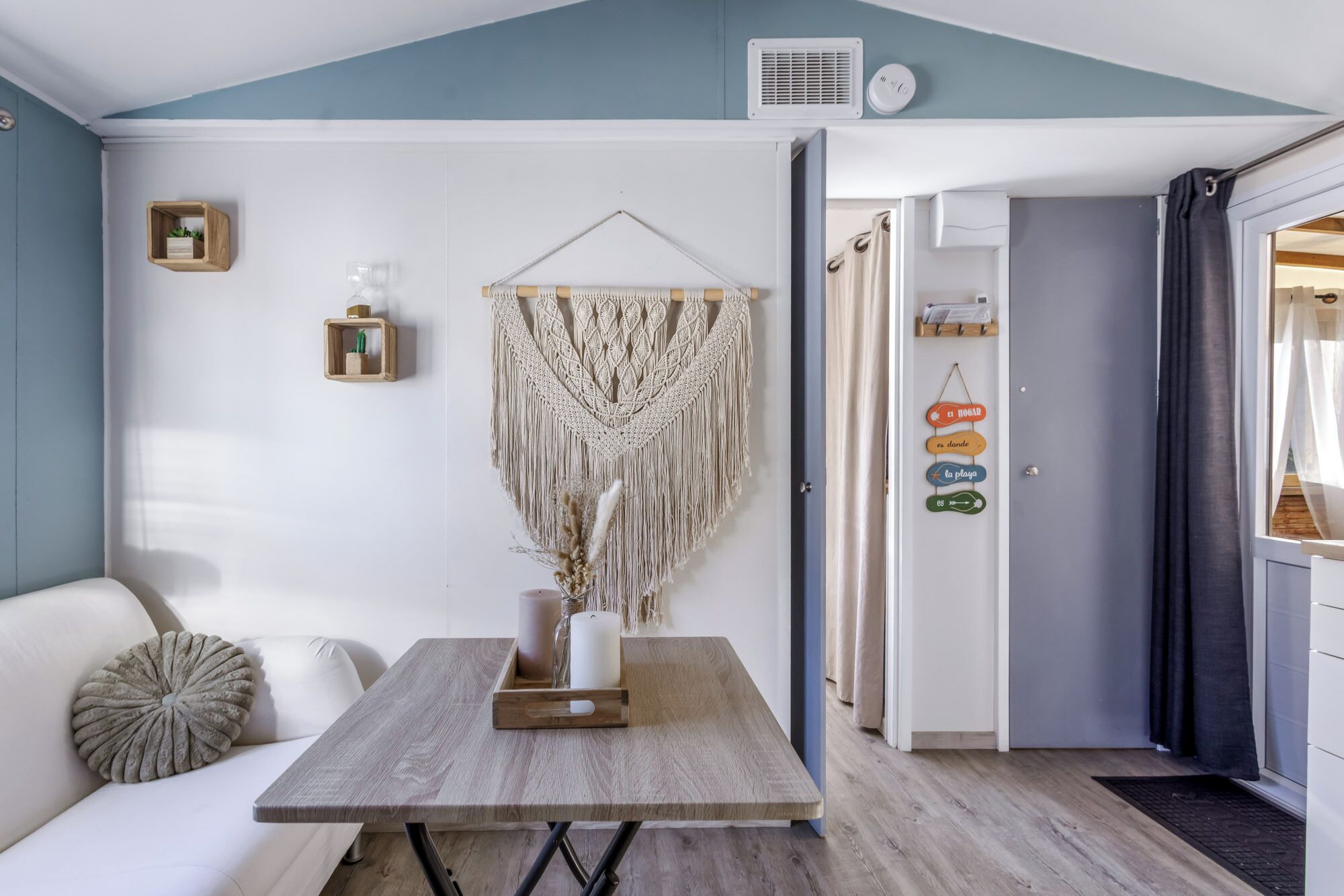 A cozy room with a wooden table, a macrame wall hanging, and a small shelf with decorative items.
