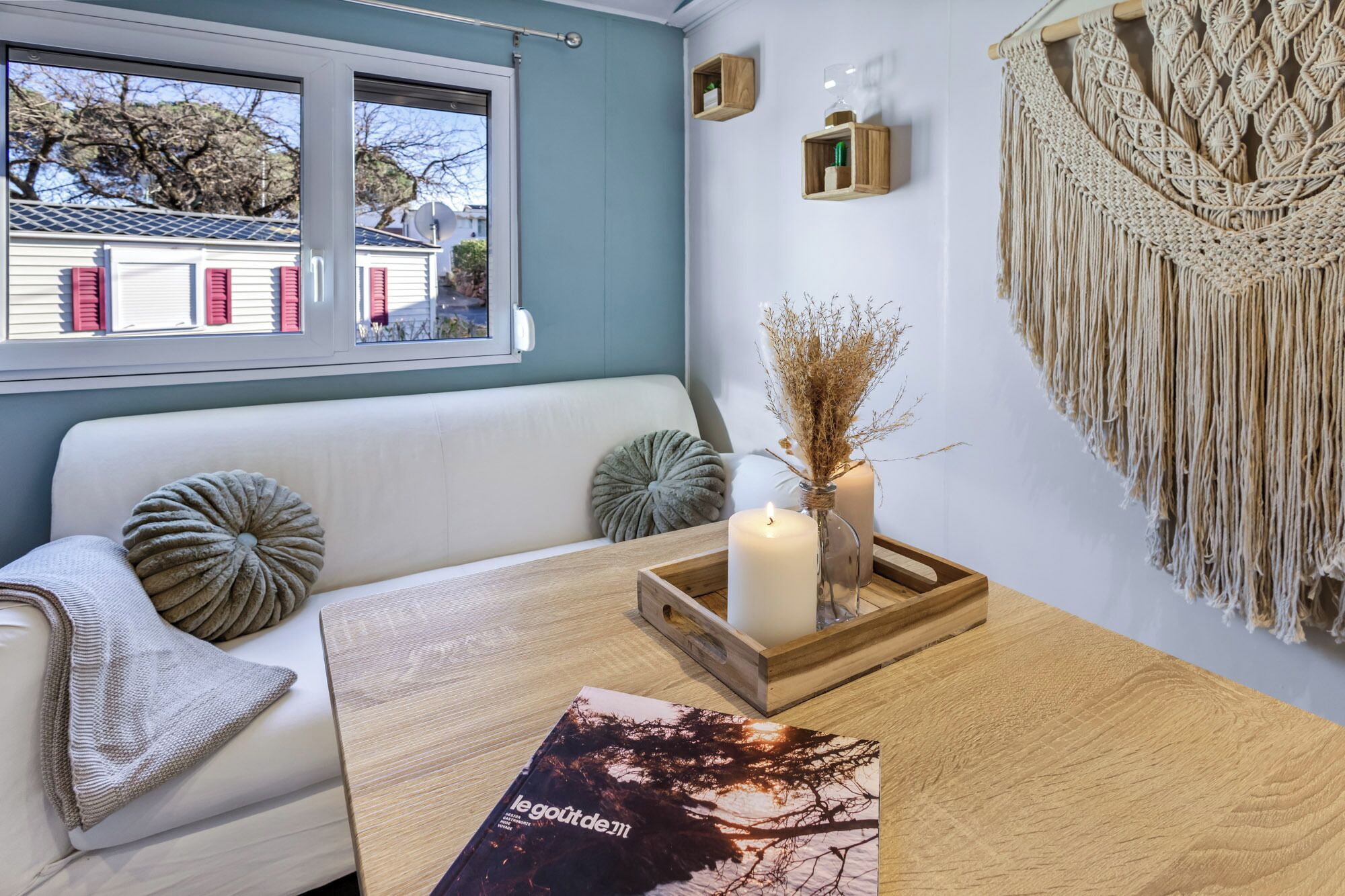 Une pièce chaleureuse avec un canapé blanc, une table d’appoint en bois, une décoration murale en macramé et une vue sur les maisons par la fenêtre.