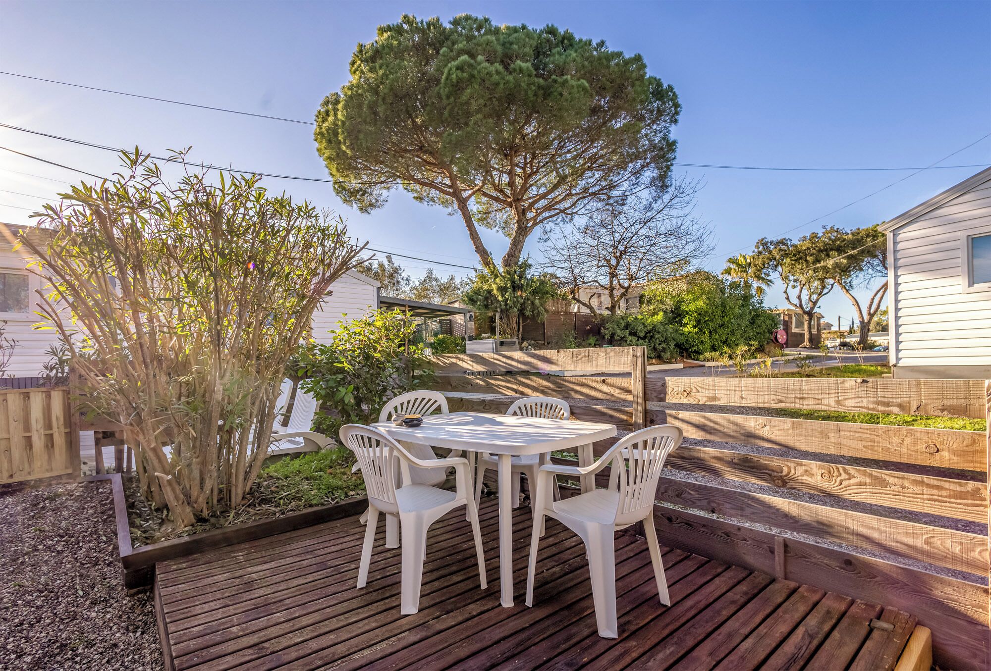 Una terraza de madera con una mesa y sillas blancas, rodeada por un jardín con un árbol grande y diversas plantas.