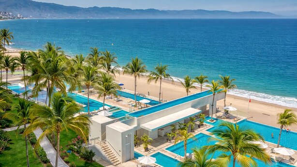 Resort com área de piscina, palmeiras e vista para a praia.